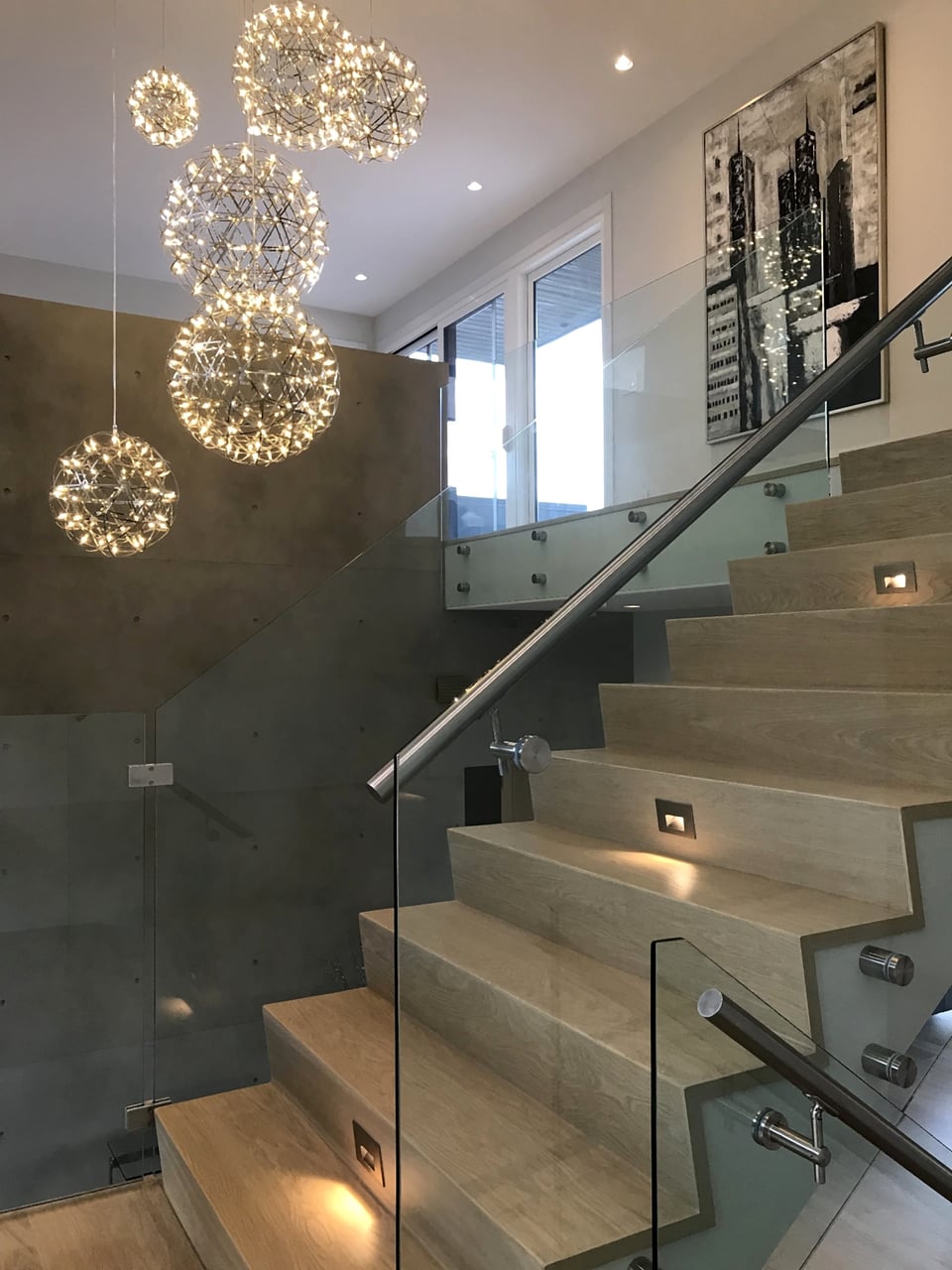 Hardwood staircase with glass railing and a cluster chandelier overhead